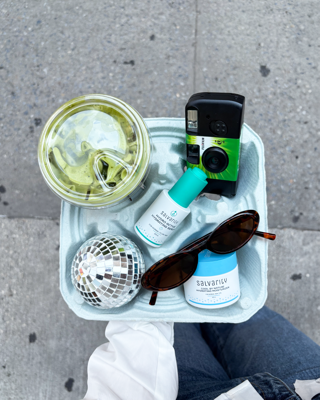 Collection of items including Salvarity produtcs bottles, sunglasses, and a camera on a tray with a concrete floor background.